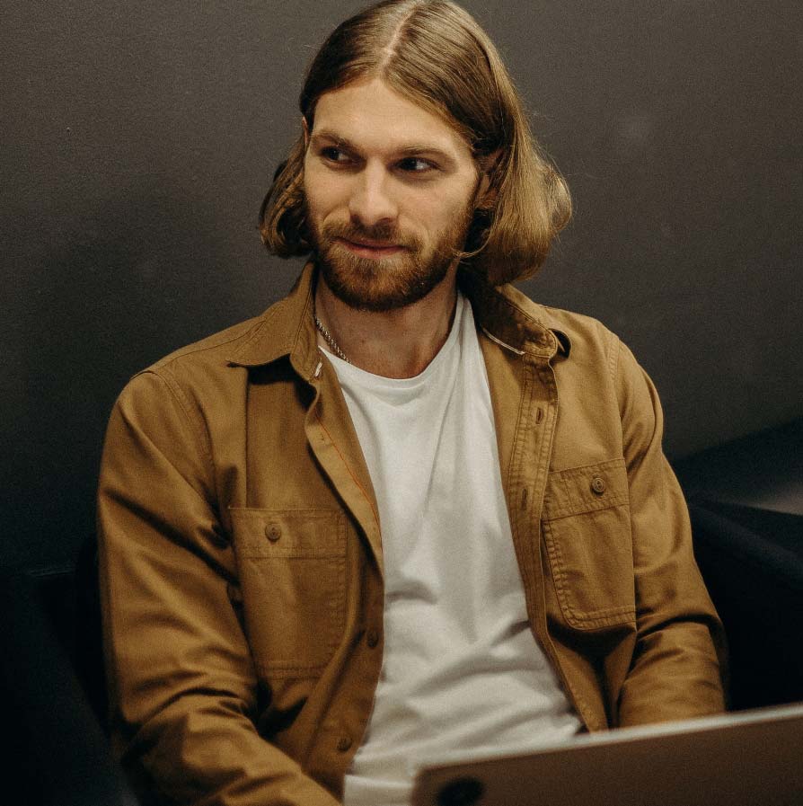 A man with long, light-brown hair wearing a brown button-down shirt over a white t-shirt.