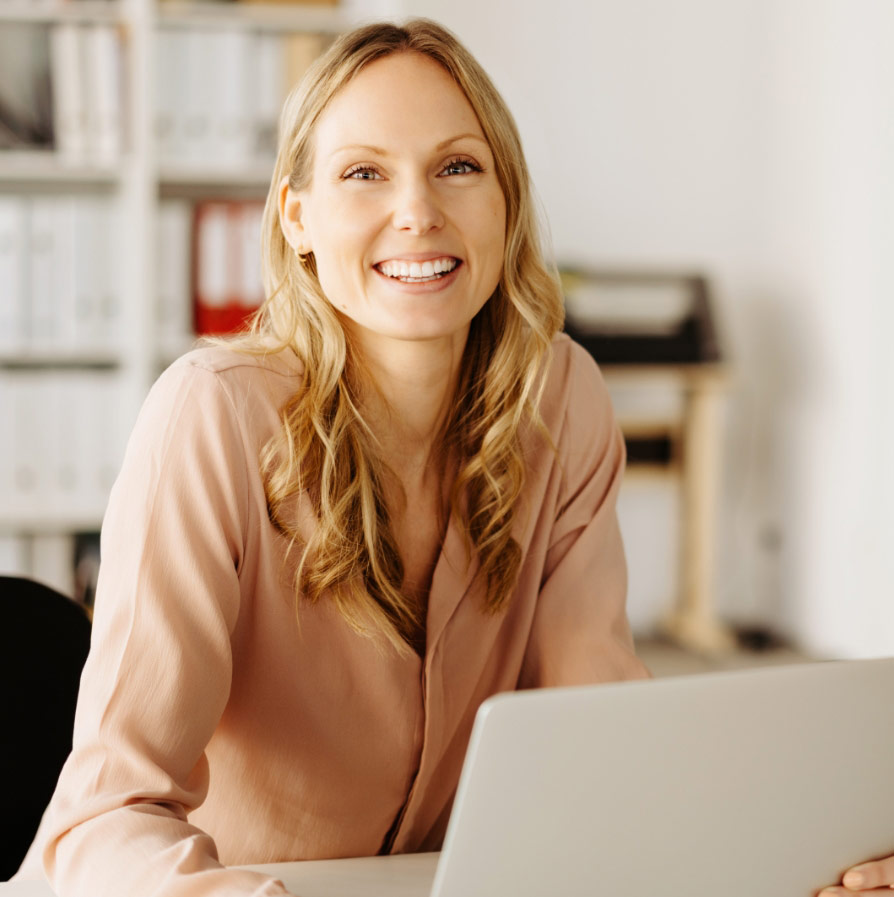 A blonde woman in a pinkish-tan blouse smiles warmly while sitting at a desk with a laptop.