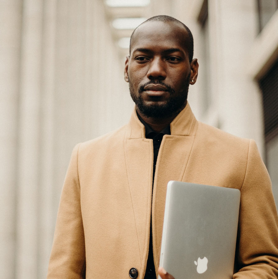 A Black man with a beard and short hair stands outdoors while holding a silver laptop.
