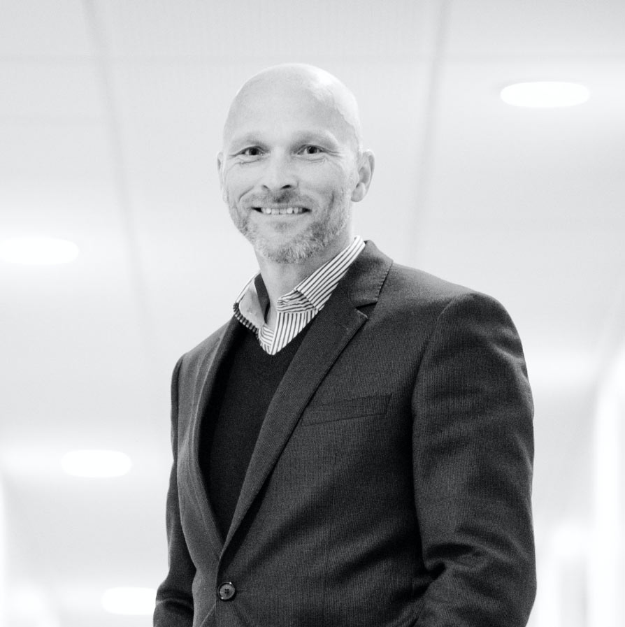 A black-and-white portrait of a smiling man wearing a suit jacket over a sweater and a striped button-down shirt.