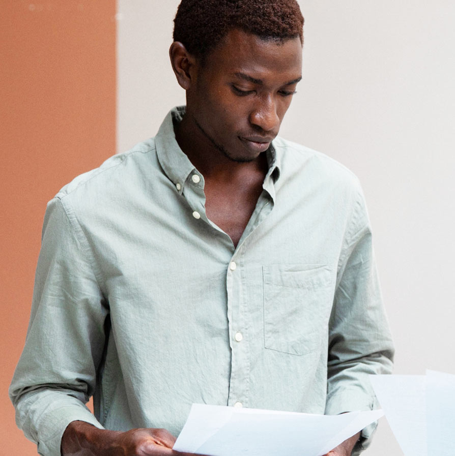A young Black man looks down while holding and reading several sheets of white paper.