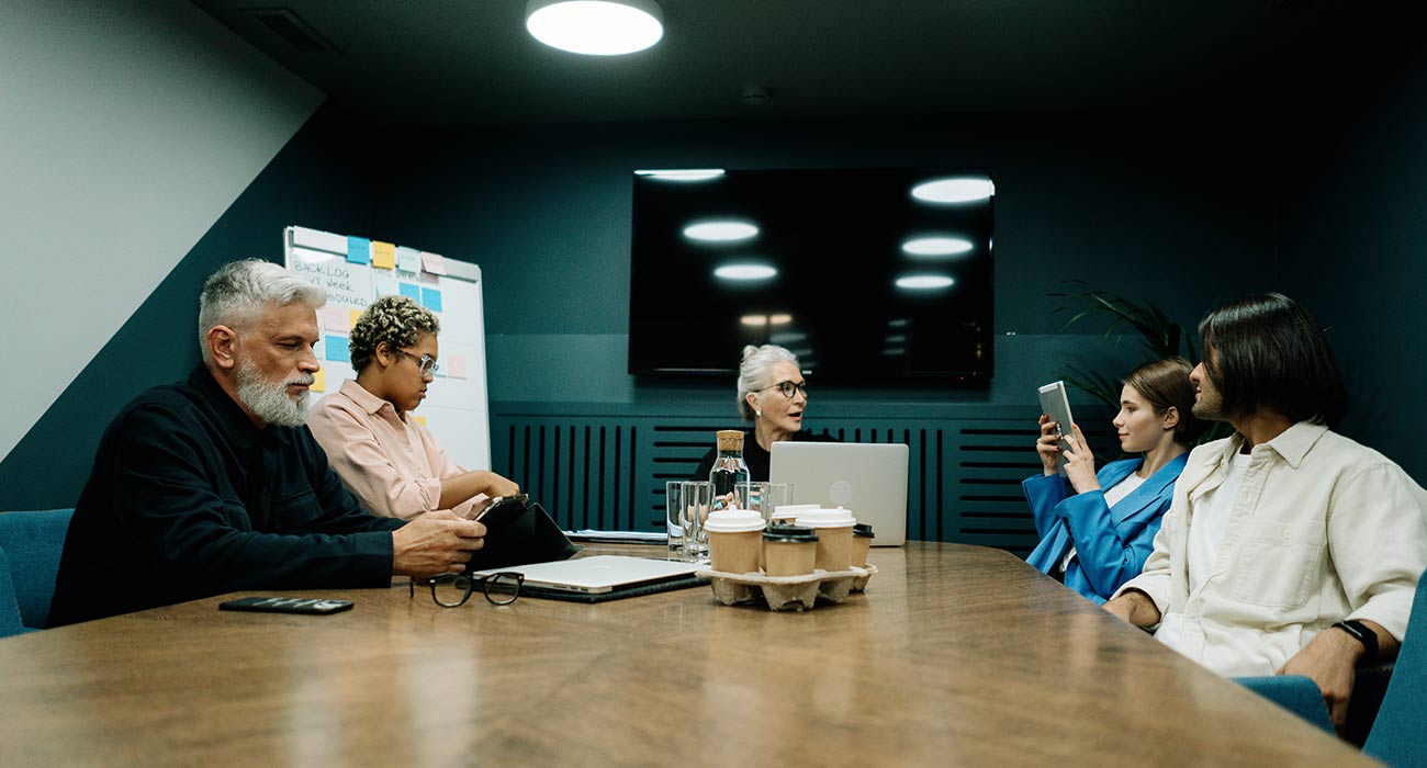 A diverse group of colleagues sits around a large wooden conference table in a modern, dimly lit meeting room.