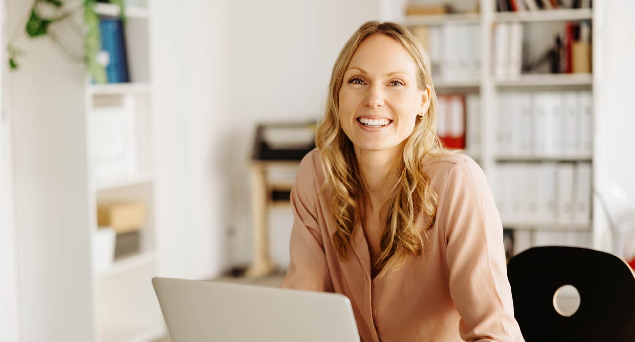 A woman with long blonde hair smiles warmly at her desk in a bright office with white bookshelves in the background.