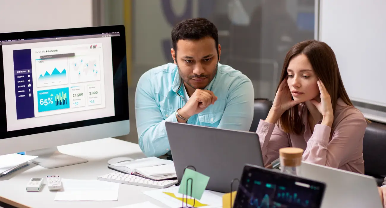 A man in a light blue shirt and a woman in a mauve blouse focused on a laptop screen in a bright office environment
