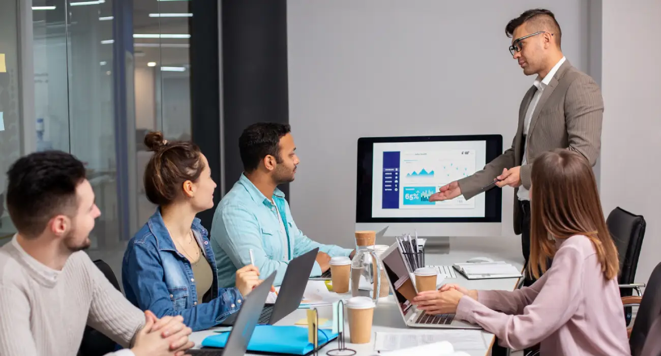 A man in a tan blazer giving a presentation to a small group of colleagues in a modern office