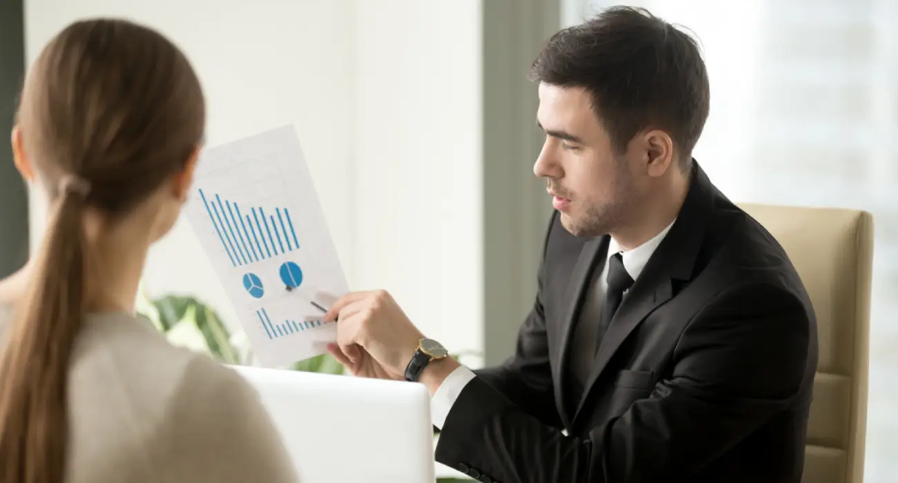 A man in a black suit pointing at a paper with blue bar and pie charts while sitting across from a woman with her hair in a ponytail.